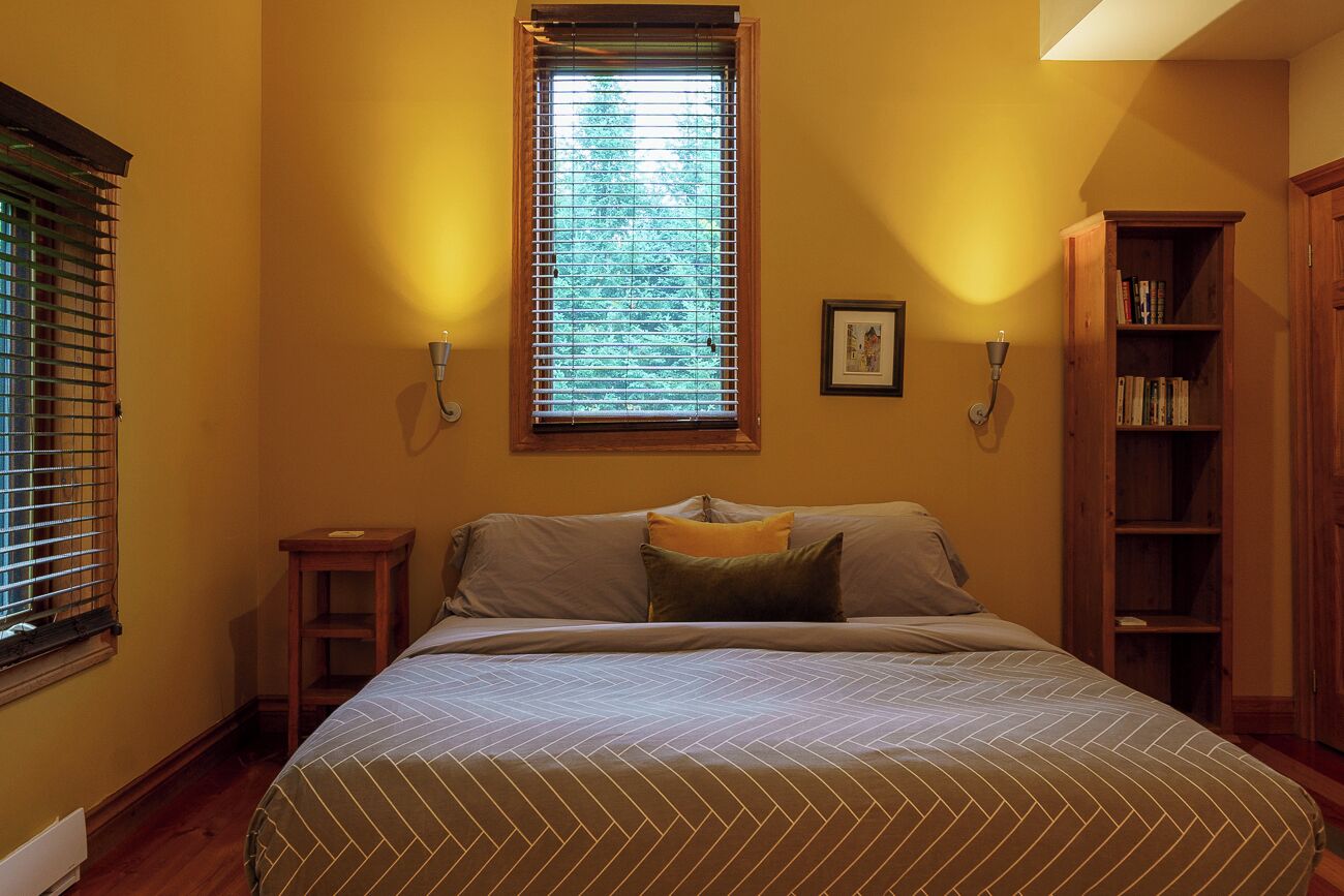 Un dormitorio con cama, ventana con persianas, mesita de noche, estantería y un cuadro en la pared.
