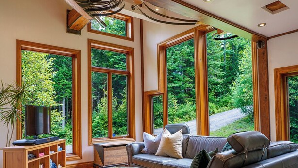 A modern living room with a leather sofa, a coffee table, and a TV on a wooden stand. Large windows offer a view of a forested area.