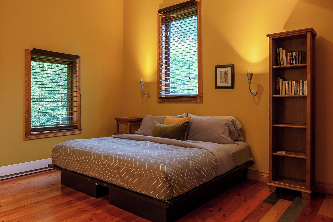 Une chambre à coucher avec un lit, deux fenêtres, une table de chevet, une bibliothèque et un tableau encadré au mur.