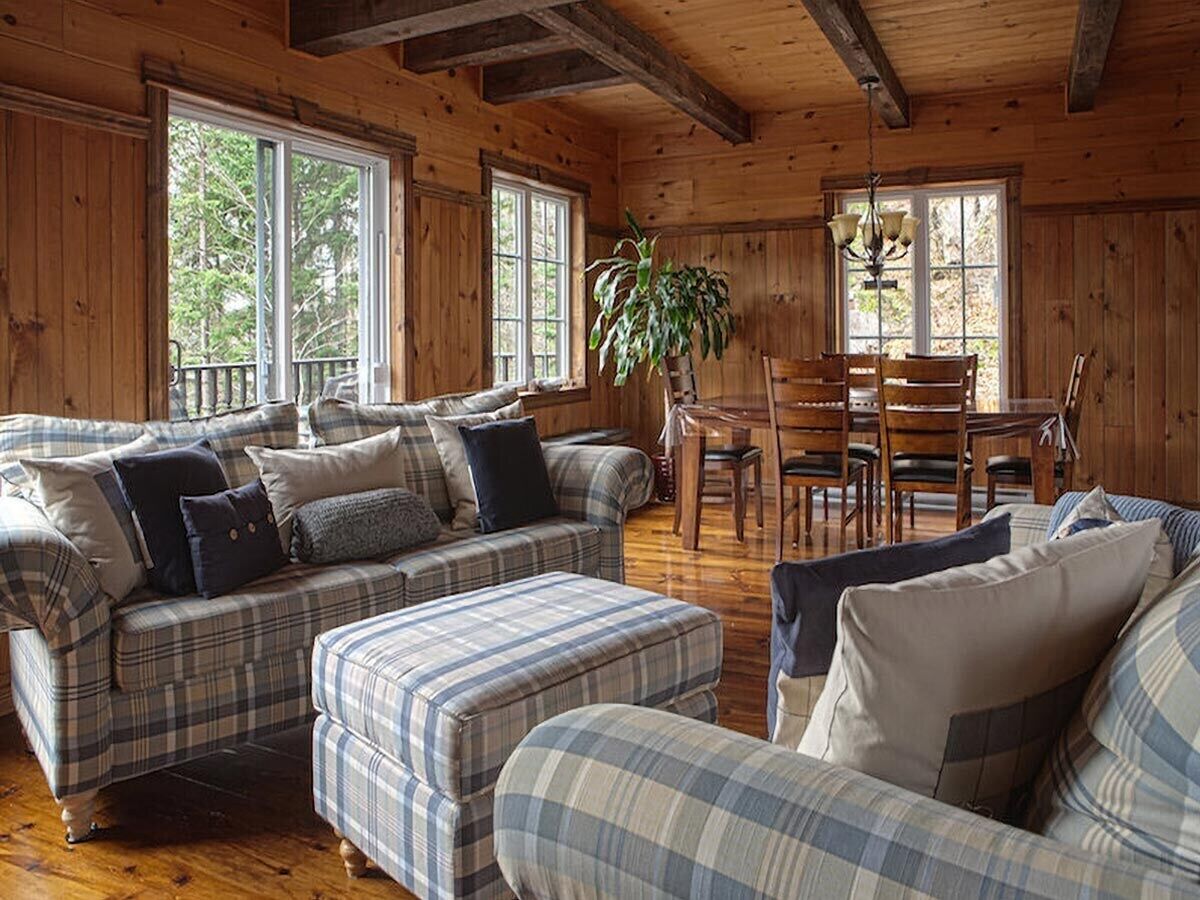 A cozy cabin interior with plaid-patterned furniture, a dining area, and large windows.