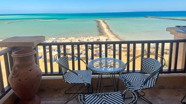 Een balkon met uitzicht op een strand, een tafel, stoelen en een decoratieve vaas.