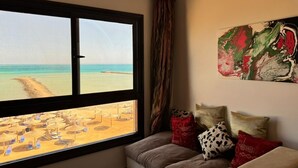 Een kamer met een groot raam dat uitkijkt op een strand, een bank met opvallende kussens, en een abstract kunstwerk aan de muur.