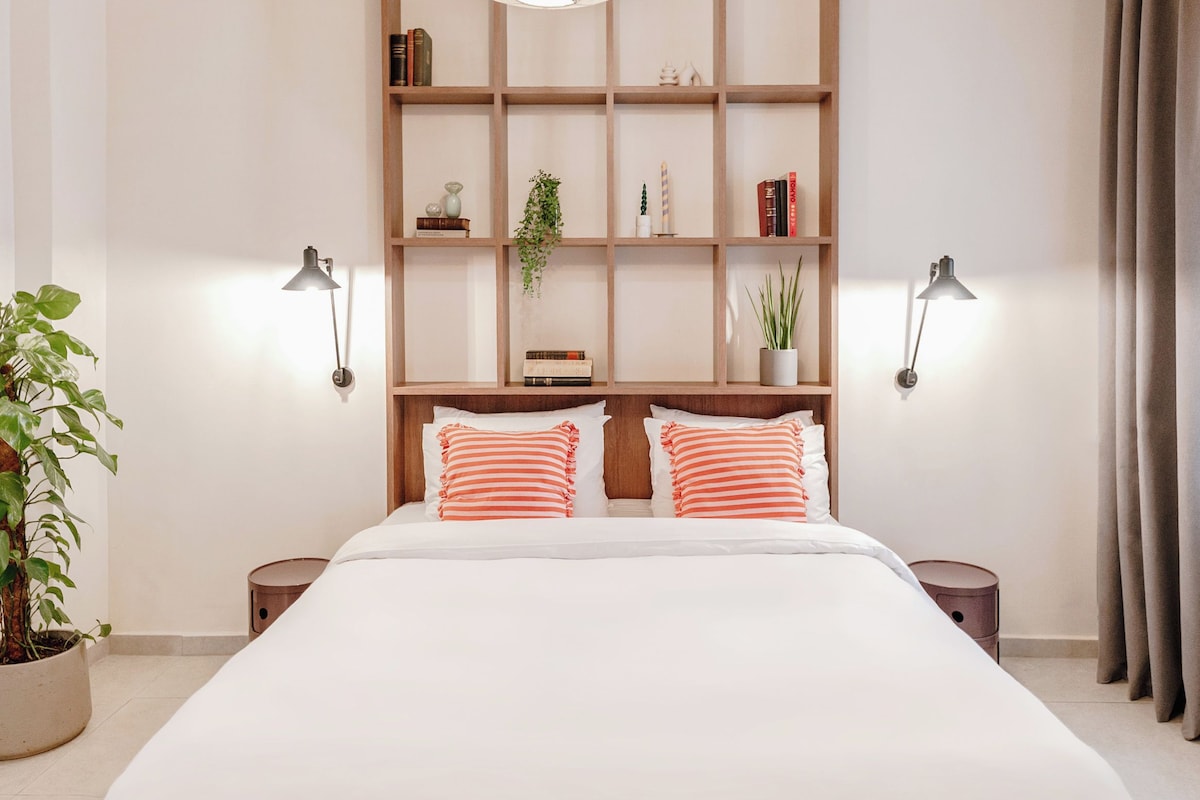 A modern bedroom with a large bed, two bedside tables, a wooden bookshelf, and hanging light fixtures.