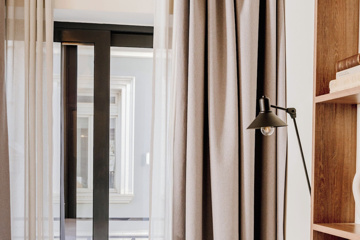 A hotel room with a bed, a green patterned pillow, a black floor lamp, and a view through a glass door.