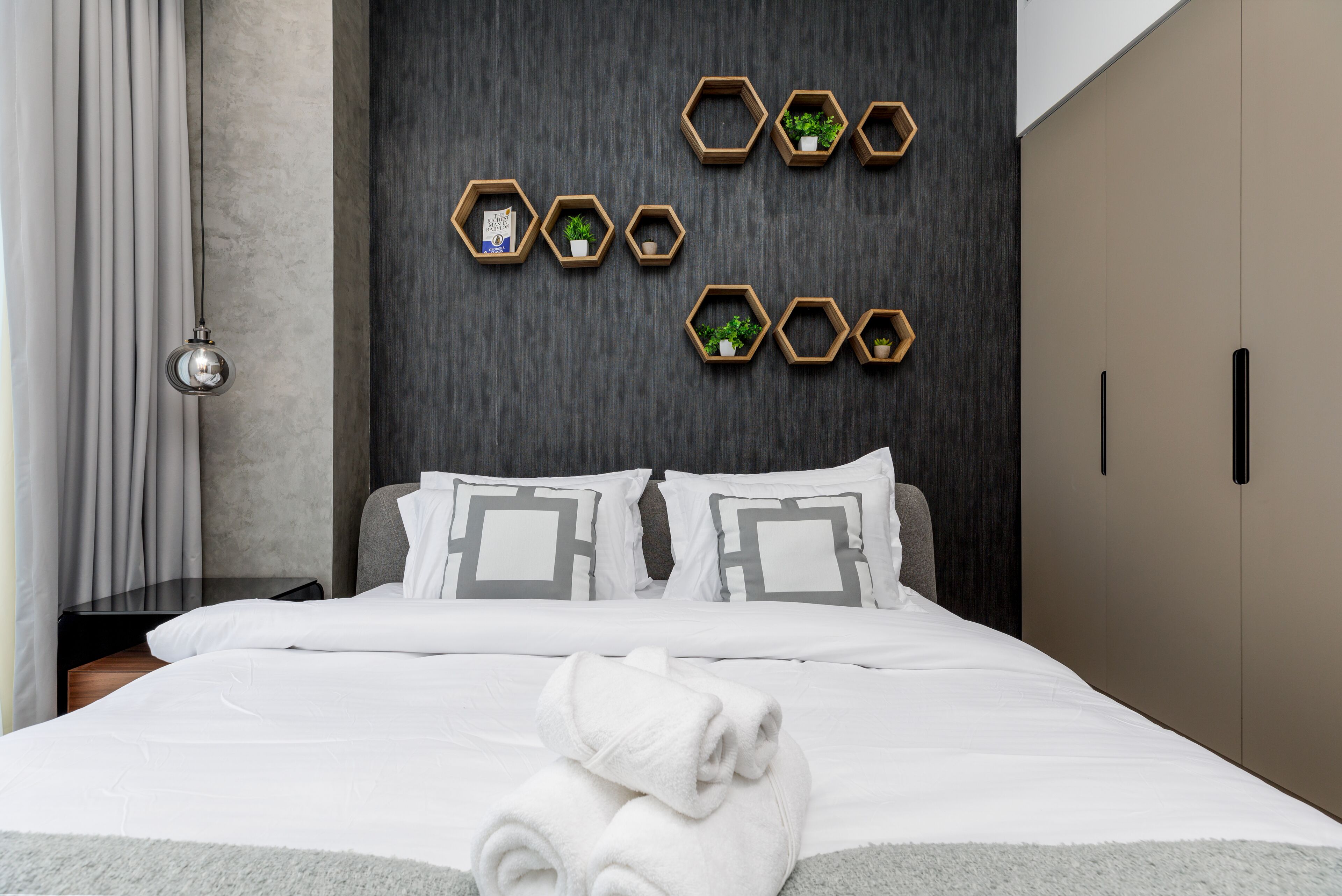 A neatly made bed with white linens and a rolled towel, a dark textured wall with hexagonal shelves holding small plants, and a modern wardrobe with black handles.