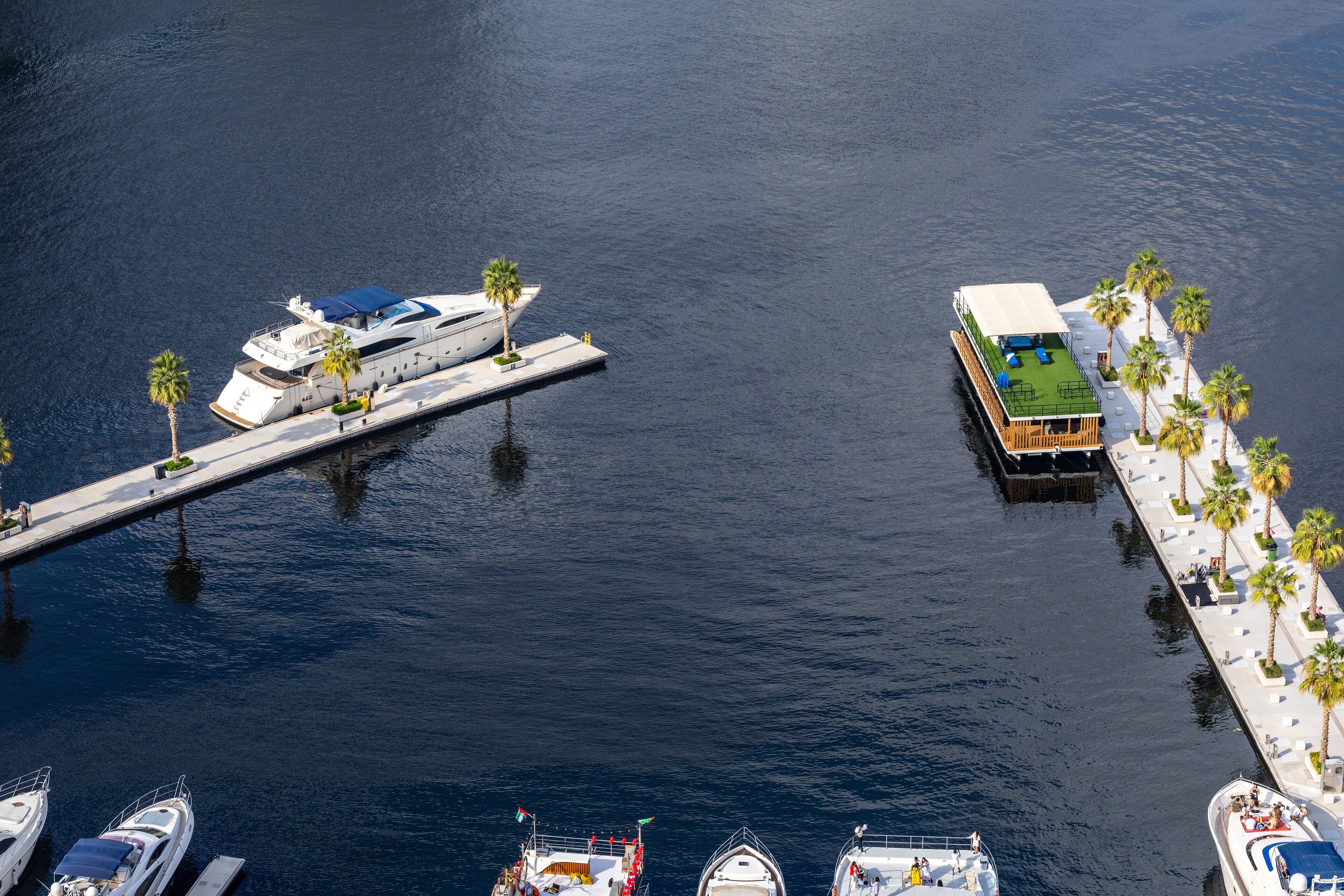 A marina with a large yacht docked at a pier, smaller boats, and a floating structure with a deck and palm trees.