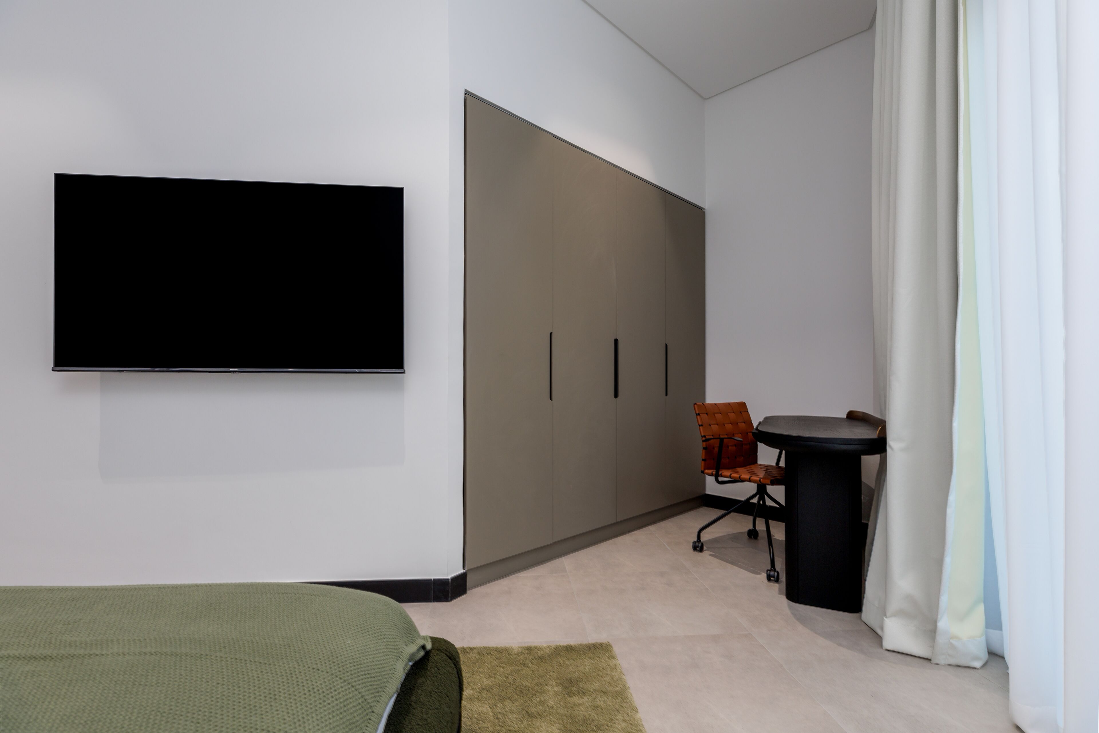 A modern hotel room with a large wardrobe, a flat-screen TV mounted on the wall, a small round table, and a green bedspread.