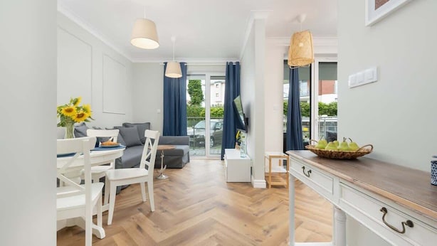 Una sala moderna con un sofá, una mesa de comedor y sillas, una zona de cocina con mostrador de madera y un amplio ventanal con cortinas.