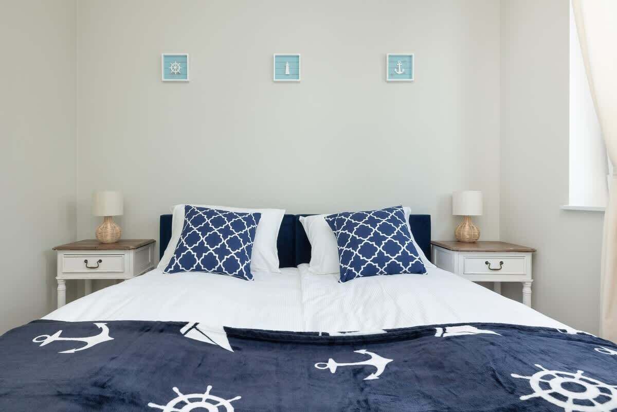 Un lit bien fait, recouvert d’une couvre-lit bleu marine et blanche aux motifs nautiques, avec deux oreillers assortis et une tête de lit blanche. On y trouve deux tables de chevet avec des lampes et de petits objets décoratifs.