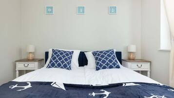 A neatly made bed with a navy blue and white nautical-themed bedspread, two matching pillows, and a white headboard. There are two bedside tables with lamps and small decorative items.