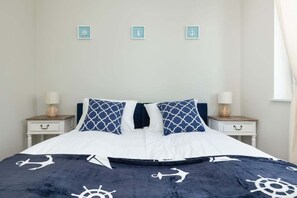 Un lit bien fait, recouvert d’une couvre-lit bleu marine et blanche aux motifs nautiques, avec deux oreillers assortis et une tête de lit blanche. On y trouve deux tables de chevet avec des lampes et de petits objets décoratifs.