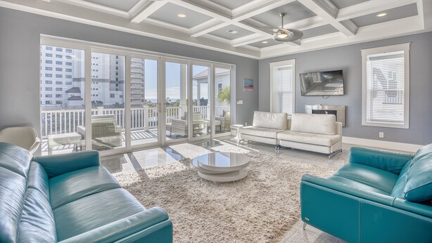 A spacious living room with a large glass door leading to an outdoor balcony, a white sofa, a teal leather couch, and a round coffee table. - Cloud IX | Luxury Home In North Beach Resort (North Myrtle Beach)