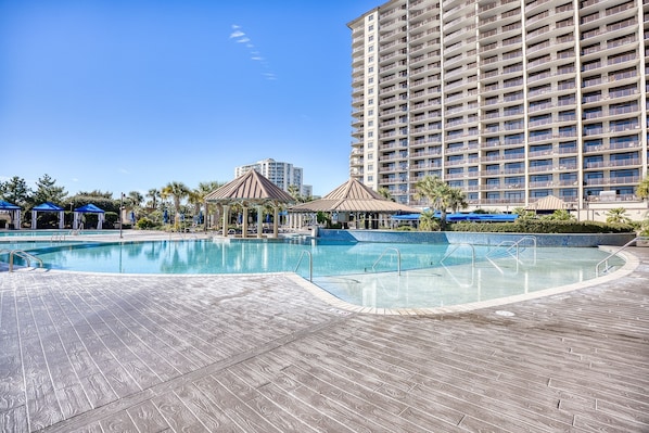 A large swimming pool with a wooden deck, surrounded by blue umbrellas and gazebos, and a tall hotel building in the background.