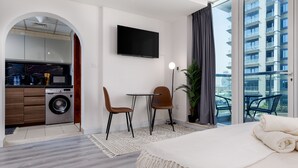 A modern hotel room with a bed, a small dining area, a TV, a washing machine, and a balcony with a view of the city. - Waves - Cozy, Fully Furnished Studio in Dubai Sports City (Dubai)
