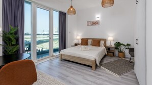 A modern hotel room with a large bed, two bedside lamps, a view of the city through the window, and a dining area with a table and chairs. - Waves - Cozy, Fully Furnished Studio in Dubai Sports City (Dubai)