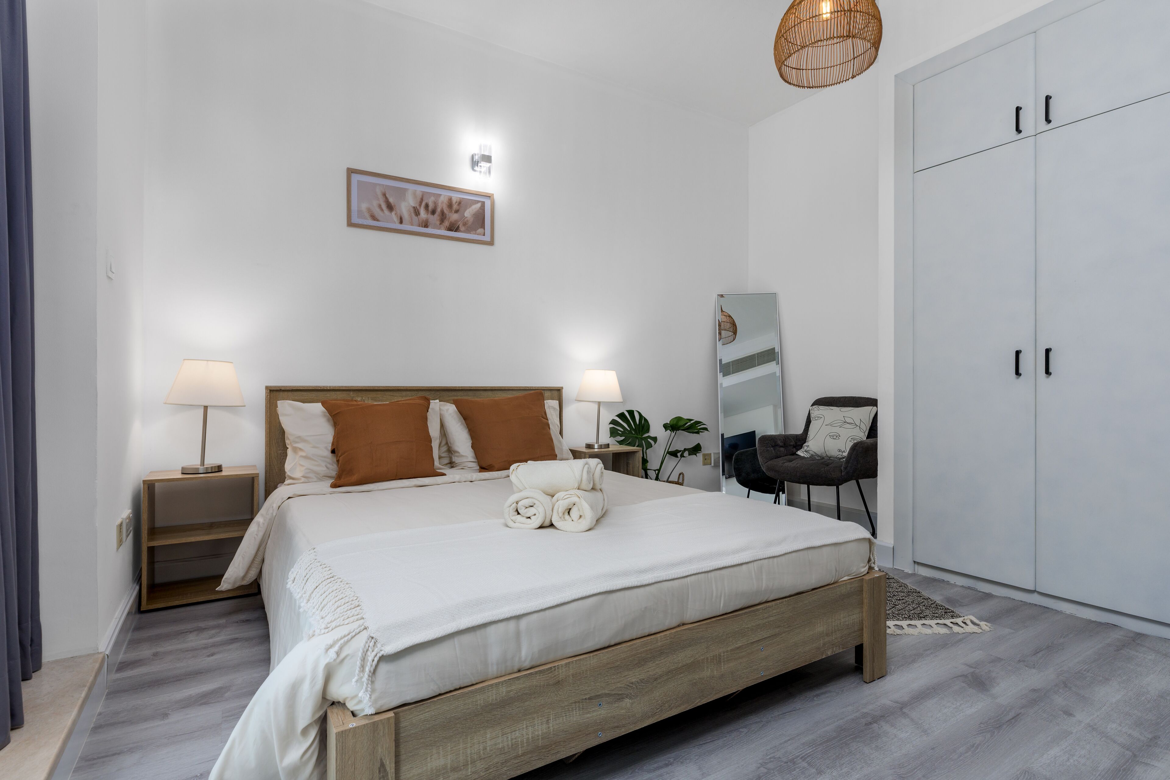 Ein ordentlich bezogenes Bett mit weißen Bettwäsche und braunen Kissen, ein Nachttisch mit Lampe, ein Kleiderschrank und ein Spiegel an der Wand.