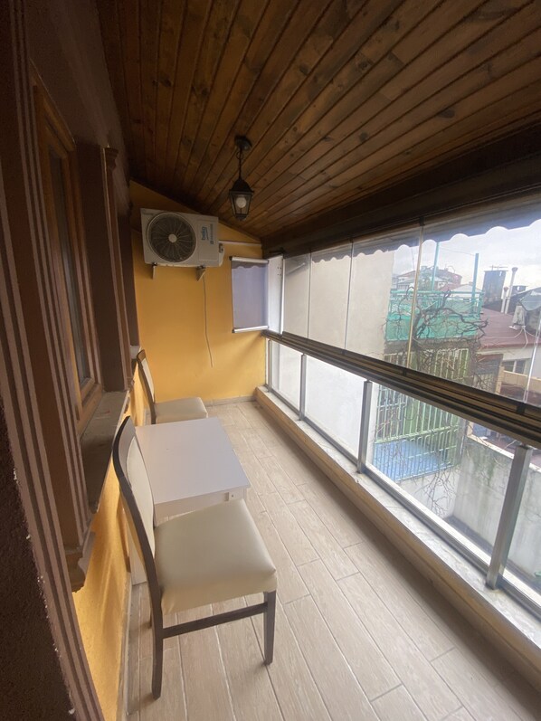 A balcony with a wooden ceiling, a table, chairs, an air conditioning unit, and a window with a view of buildings.