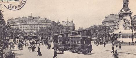A historical Parisian street scene with horse-drawn carriages, a double-decker bus, and a statue.