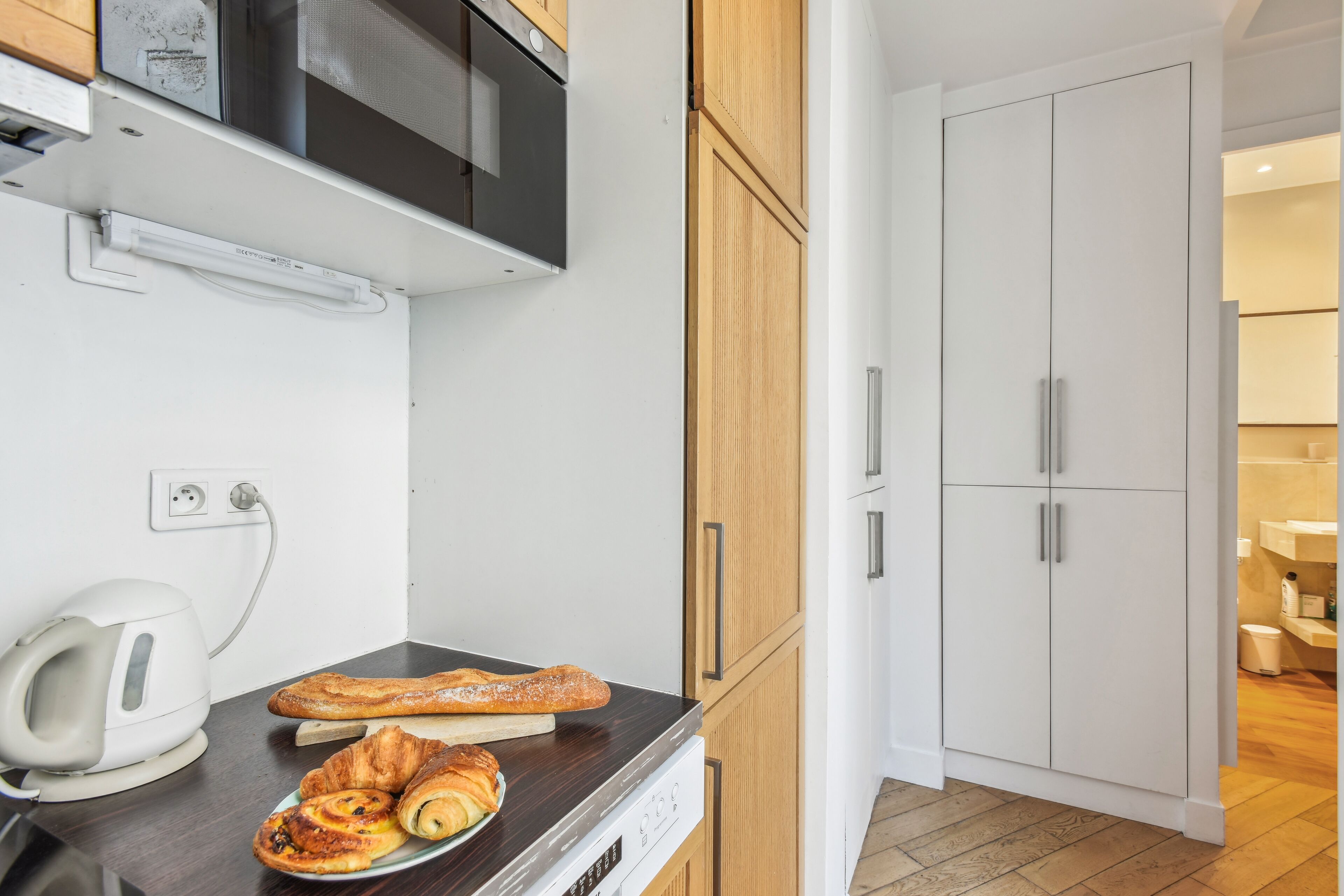 A kitchenette with a microwave, toaster, and a plate of pastries. There is a wooden cabinet and a white door in the background.