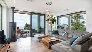 A modern living room with a large sofa, a wooden coffee table, and a fireplace. There is a balcony with a view of the ocean and palm trees.