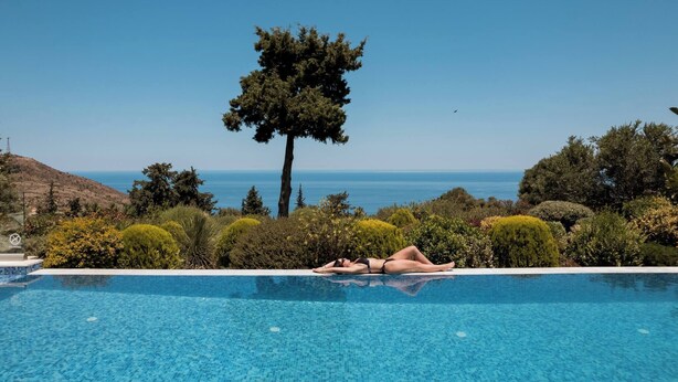 A woman lying on a poolside lounge chair with a clear view of the ocean and a tree in the background.