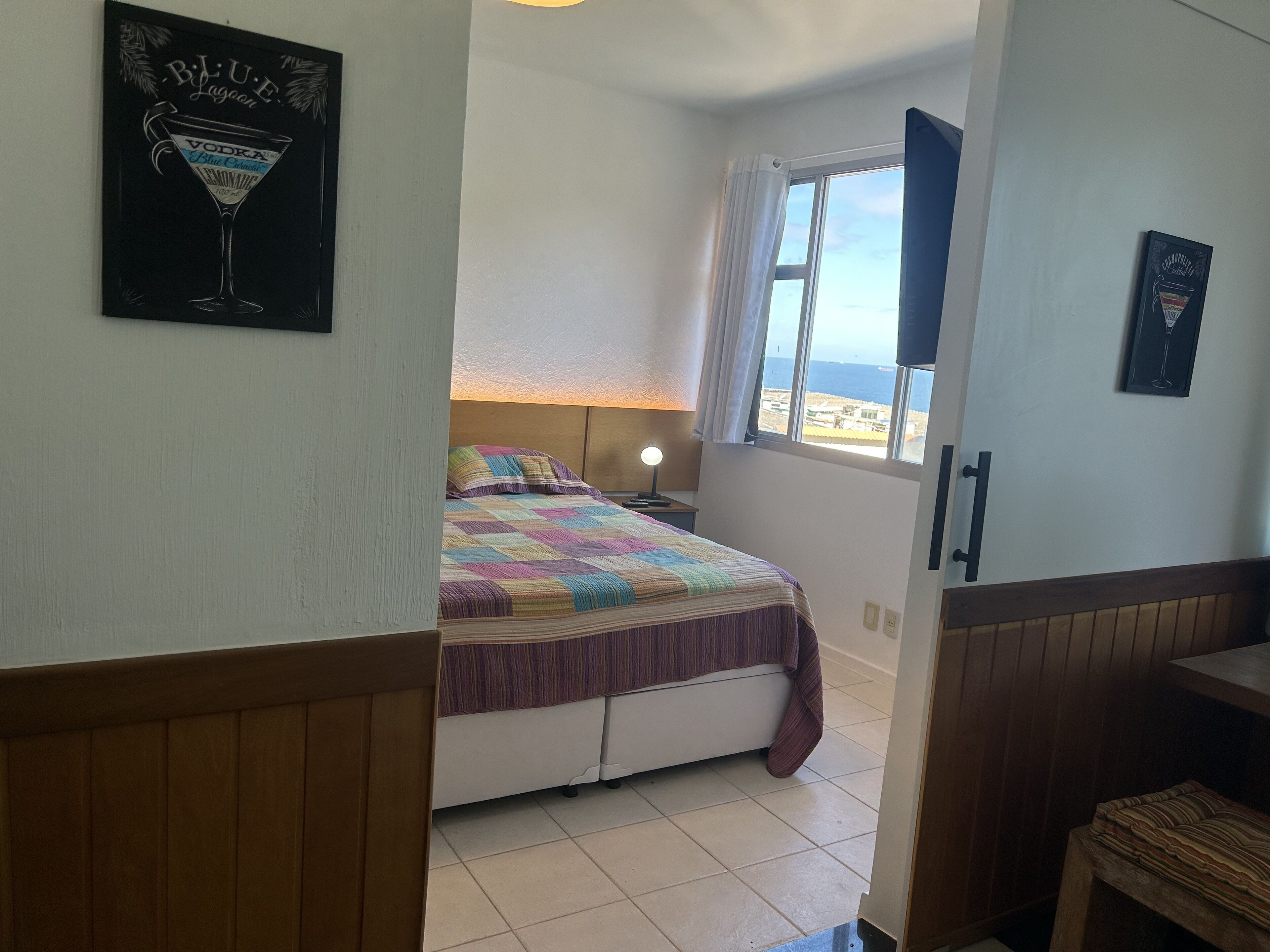 A bedroom with a bed, a nightstand, a lamp, a window with a view, and a door leading to another room.