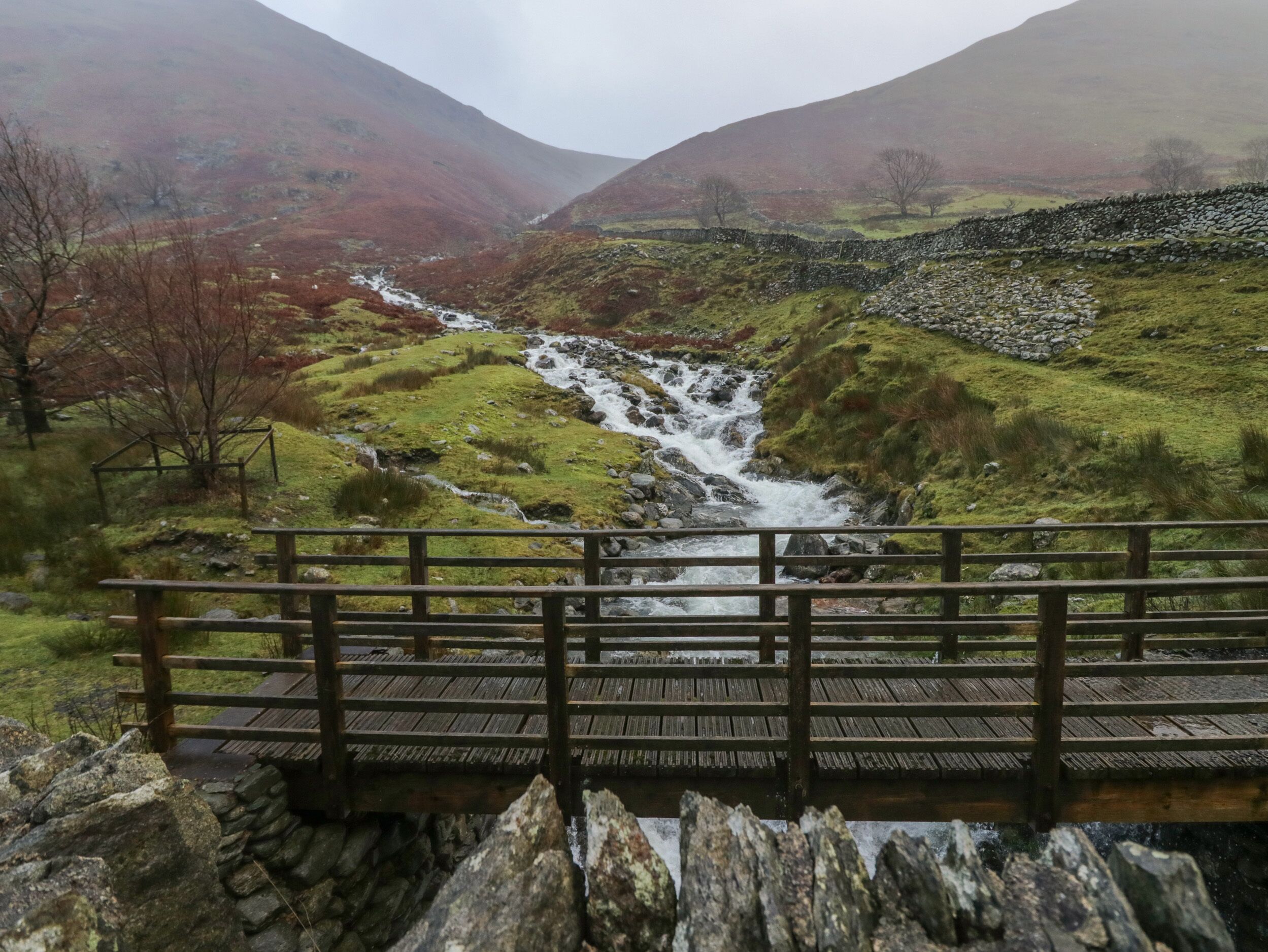 A wooden bridge over a rocky stream with a mountainous backdrop.
