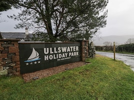 Placa para o Ullswater Holiday Park com logotipo de um veleiro, localizada na entrada de um parque com muro de pedra e área gramada.