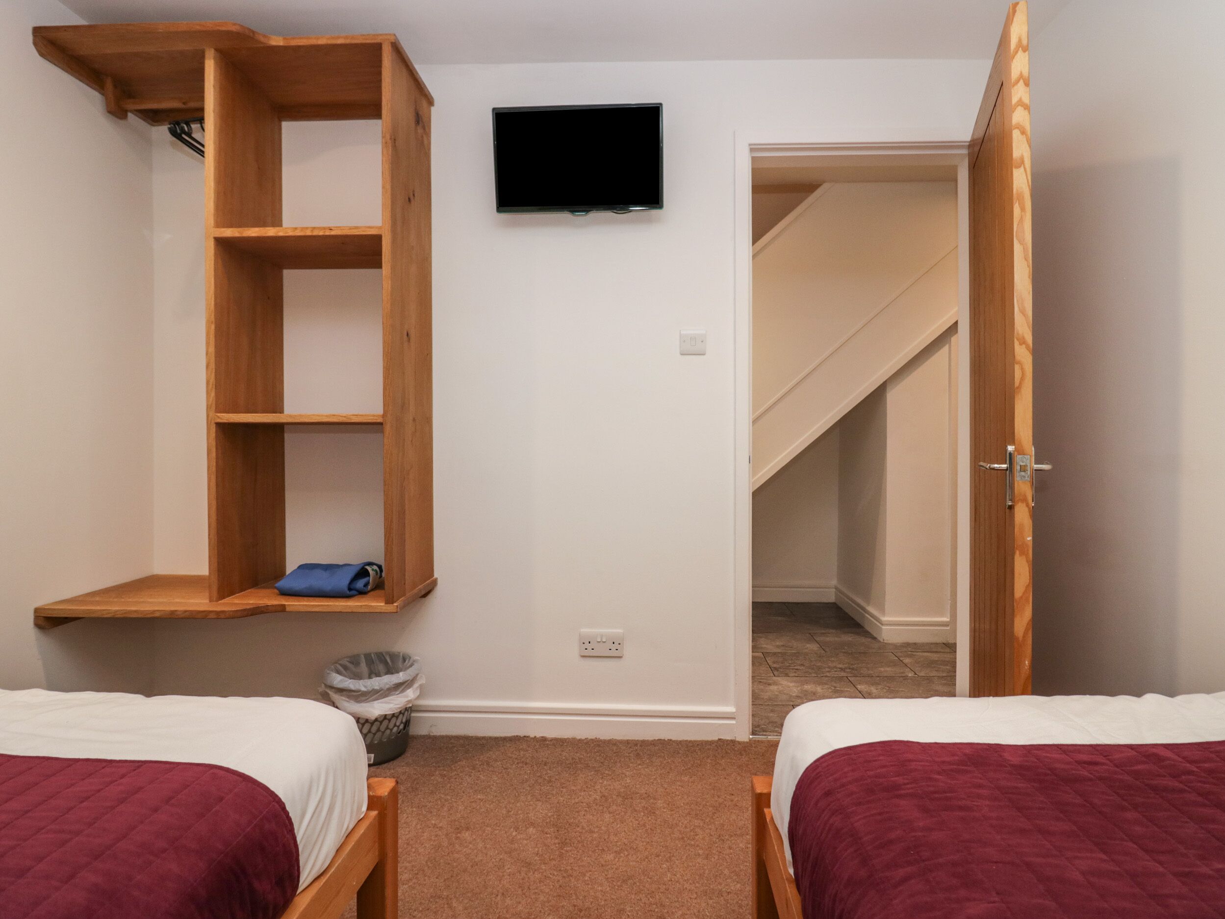 A bedroom with two beds, a TV mounted on the wall, a wooden shelf, and a door leading to a staircase.