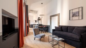 Een moderne woonkamer met een donkerblauwe bank, een grijze fauteuil en een salontafel. Links staat een tv op een meubel en er is een eethoek op de achtergrond.