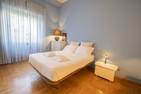 Una cama bien hecha con sábanas y toallas blancas, una mesita de noche con lámpara, y una ventana con cortinas traslúcidas.