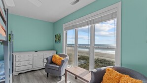 A room with teal walls, a large window with a beach view, a white dresser, two gray armchairs with yellow pillows, and a wooden coffee table. - 5 Bedroom Oceanfront home with private pool! (Oak Island)