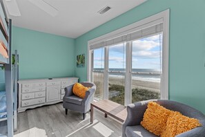 Quarto com paredes verde-água, grande janela vista para a praia, cômoda branca, duas poltronas cinzas com almofadas amarelas e mesa de centro de madeira.