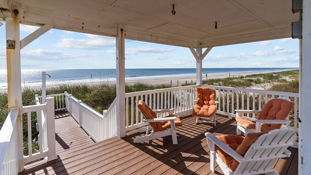 A covered deck with white railings and wooden flooring, overlooking a beach with a clear view of the ocean and sandy shore. - 5 Bedroom Oceanfront home with private pool! (Oak Island)