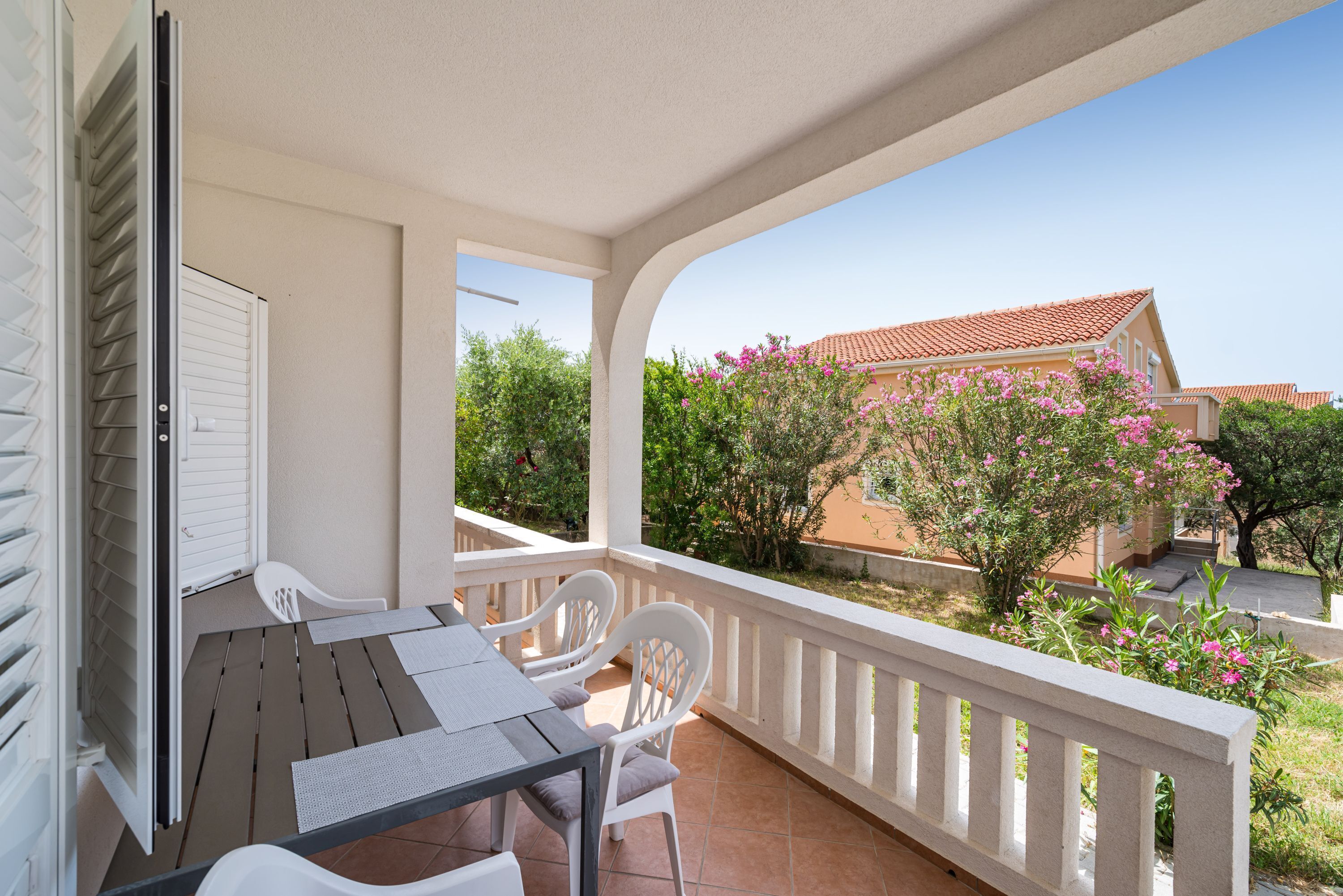A balcony with a table and chairs, a white railing, and a view of a building with a red roof and pink flowers.