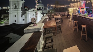 Un bar en la azotea con mesas y sillas de madera, luces colgantes y vista a un edificio colonial blanco al fondo.