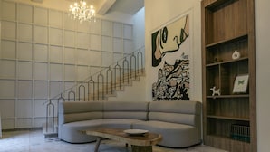 Una sala de estar moderna con piso de piedra, un sofá grande, una mesa de centro de madera, una araña, una escalera y una obra de arte abstracta de gran tamaño en la pared.