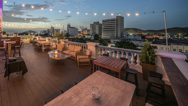 Una terraza al aire libre con mesas y sillas de madera, luces colgantes y vistas a la ciudad al fondo.