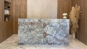 Un mostrador de recepción con encimera de mármol, paneles de madera en las paredes, una estantería con premios y una planta decorativa alta.