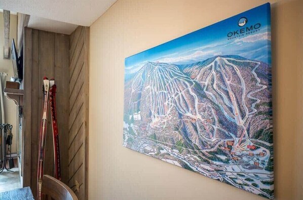 A large painting of a mountain resort with a ski lift, located in a room with wooden walls and ski equipment.