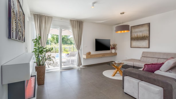 A modern living room with a grey sofa, a flat-screen TV mounted on the wall, a potted plant, and a sliding glass door leading to an outdoor area.