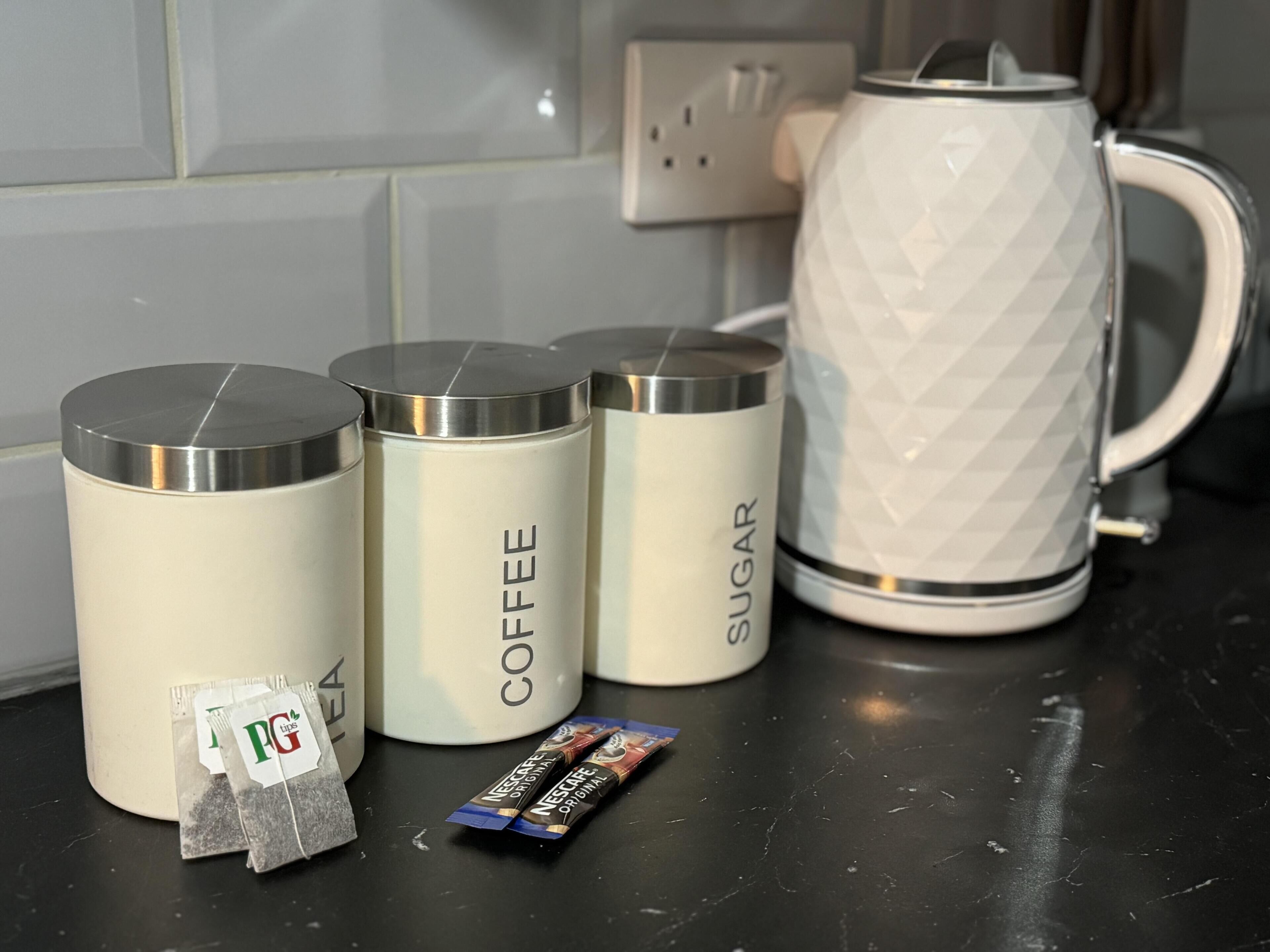 Four stainless steel containers labeled 'TEA,' 'COFFEE,' and 'SUGAR' on a kitchen counter, with a white kettle and a packet of tea and coffee in front.