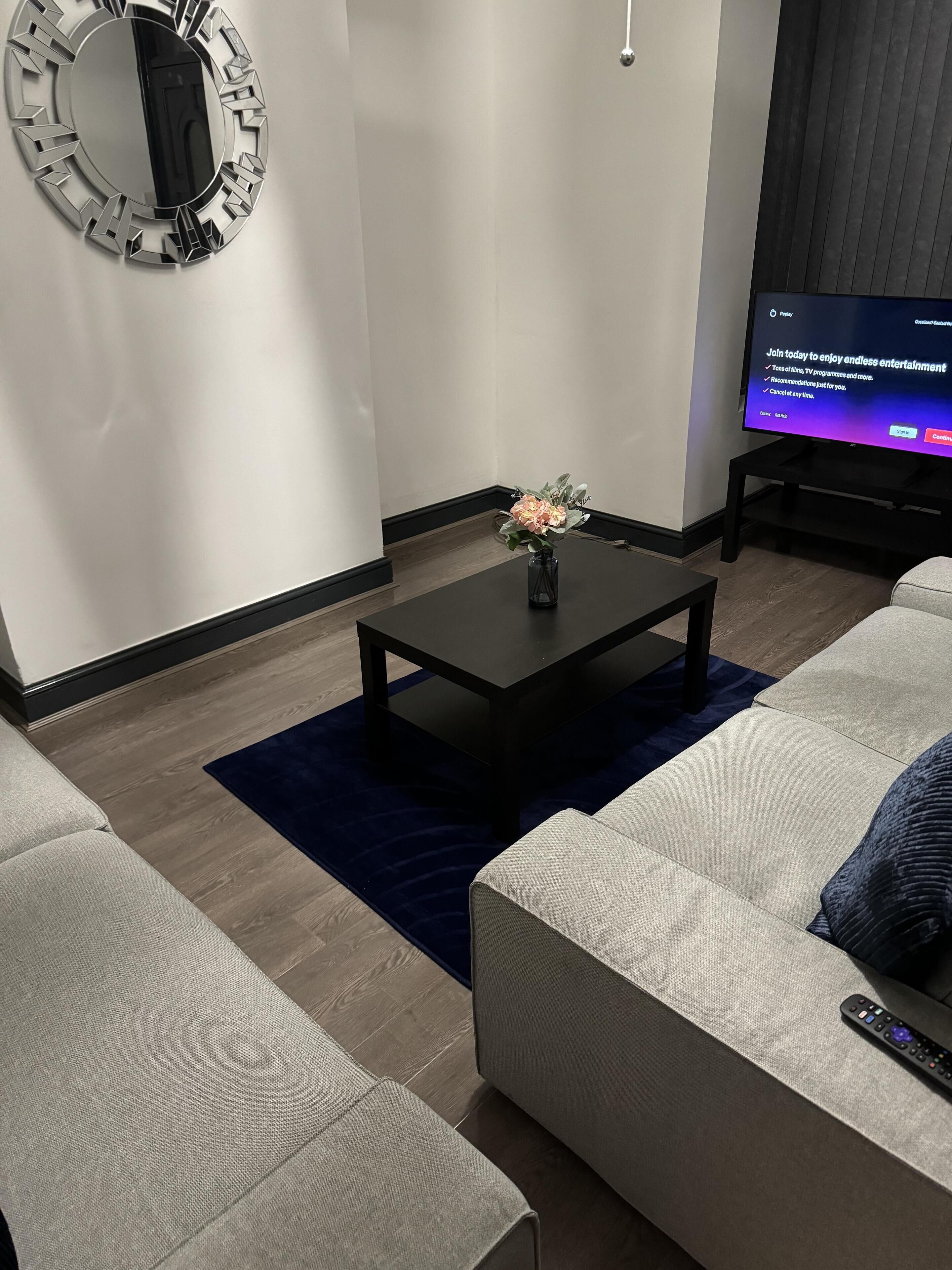 A modern living room with a black coffee table, a vase of flowers, a grey sofa, and a television displaying a sign.