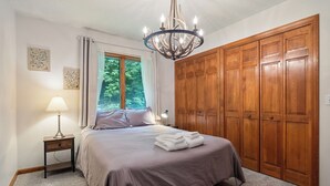 Een slaapkamer met een bed, een nachtkastje, een lamp, een kroonluchter, een raam met gordijnen en houten kasten.