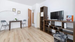 A modern living room with a wooden floor, a TV on a wooden stand, a dining table with chairs, and a bookshelf.