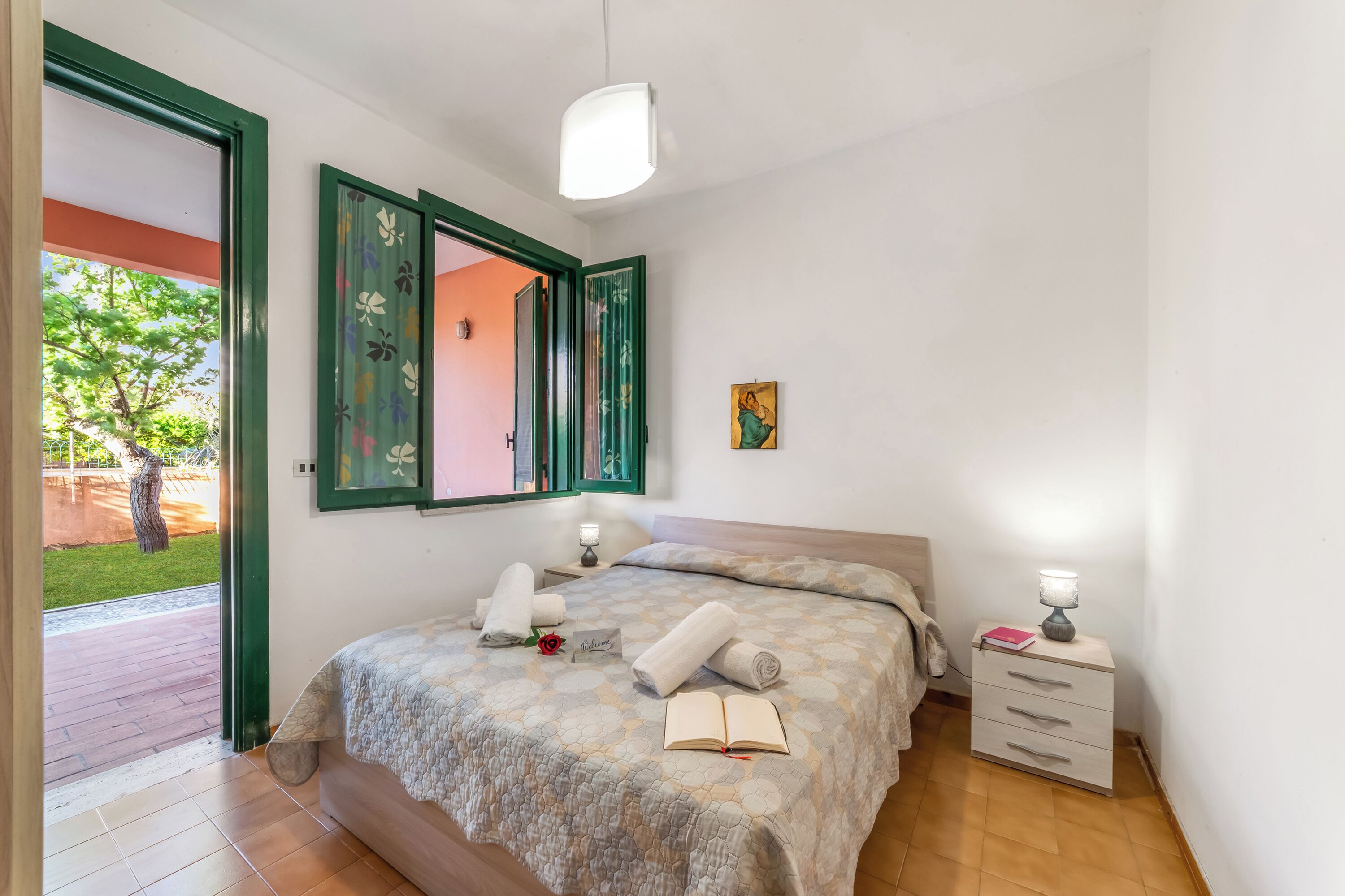 A bedroom with a bed, a bedside table, a nightstand, a door, and a view of the outside.
