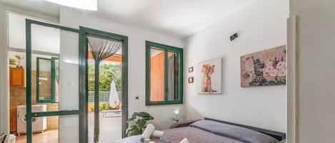 A bedroom with a bed, a window, a painting, and a view of the outside.