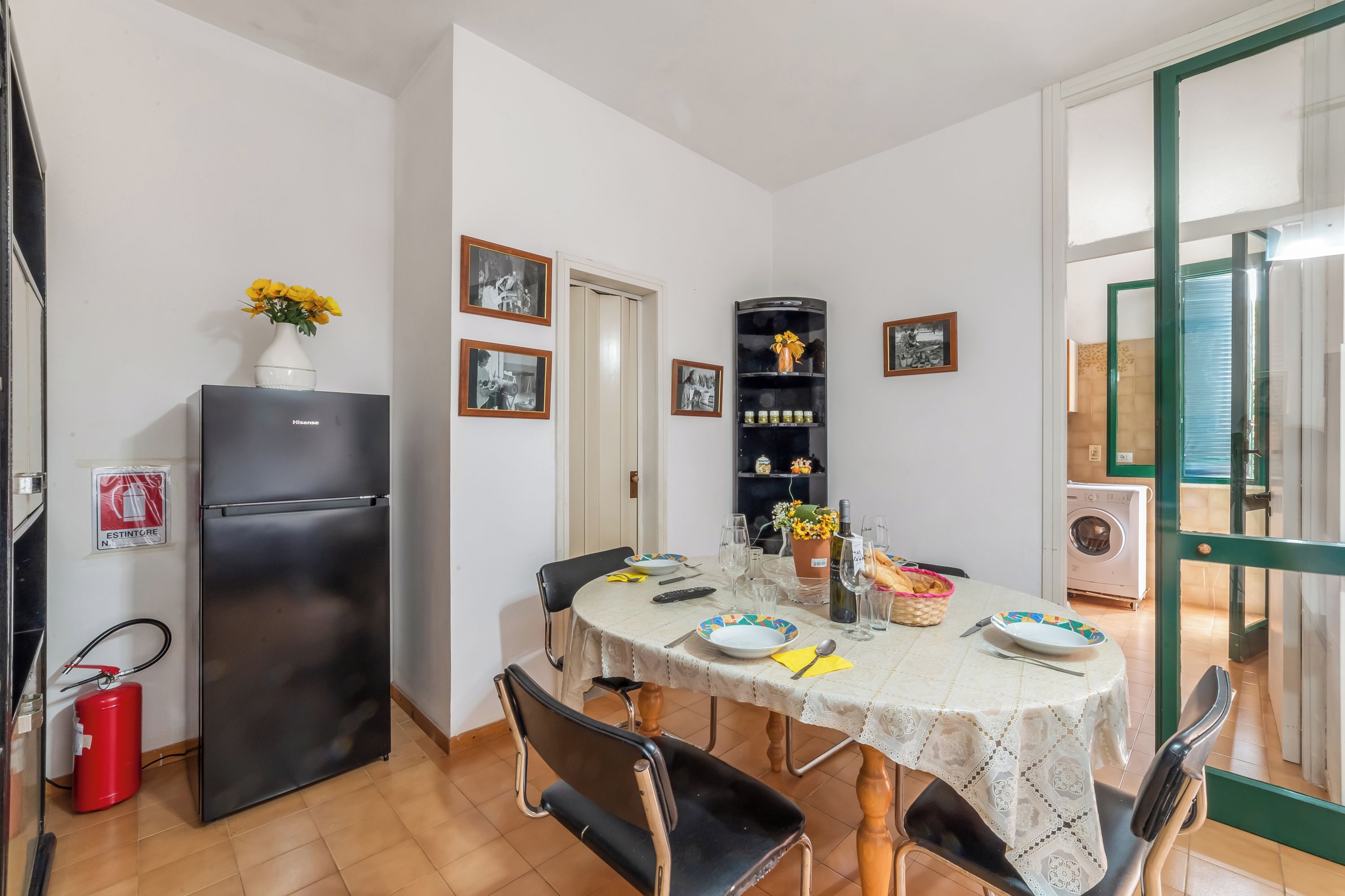 A dining area with a table set for four, a black refrigerator, a fire extinguisher, and a shelf with decorative items.