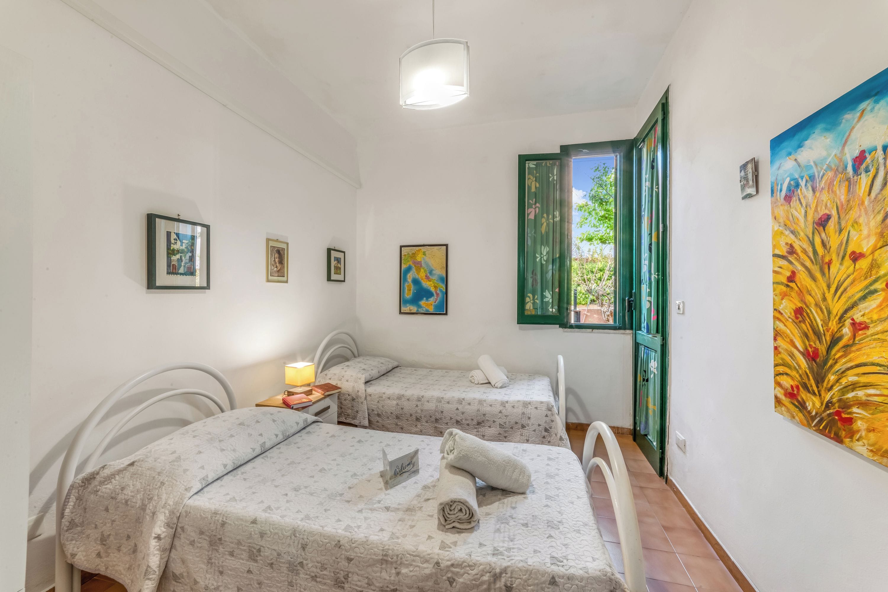 Une chambre avec deux lits, une petite table avec une lampe, un tableau au mur et une fenêtre avec des volets verts.