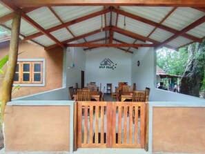 A covered outdoor seating area with wooden tables and chairs, a metal roof, and a wall with a logo. - Wilpattu wild villa (Pahala Maragahawewa)